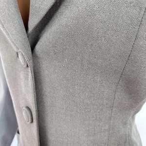 Puede incluir: Primer plano de una blazer gris claro con solapa de muesca y tres botones a juego. La tela tiene un sutil tejido diagonal, y la blazer parece estar hecha a medida. El estilo general es cl&aacute;sico y profesional.