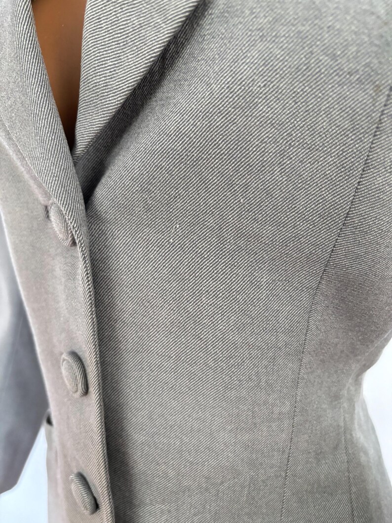 Puede incluir: Primer plano de una blazer gris claro con solapa de muesca y tres botones a juego. La tela tiene un sutil tejido diagonal, y la blazer parece estar hecha a medida. El estilo general es cl&aacute;sico y profesional.