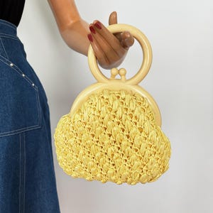 May include: A vintage yellow raffia handbag with a cream-colored handle and clasp. The bag has a rounded shape and a woven texture. The handle is a circular design. The bag is held by a mannequin.