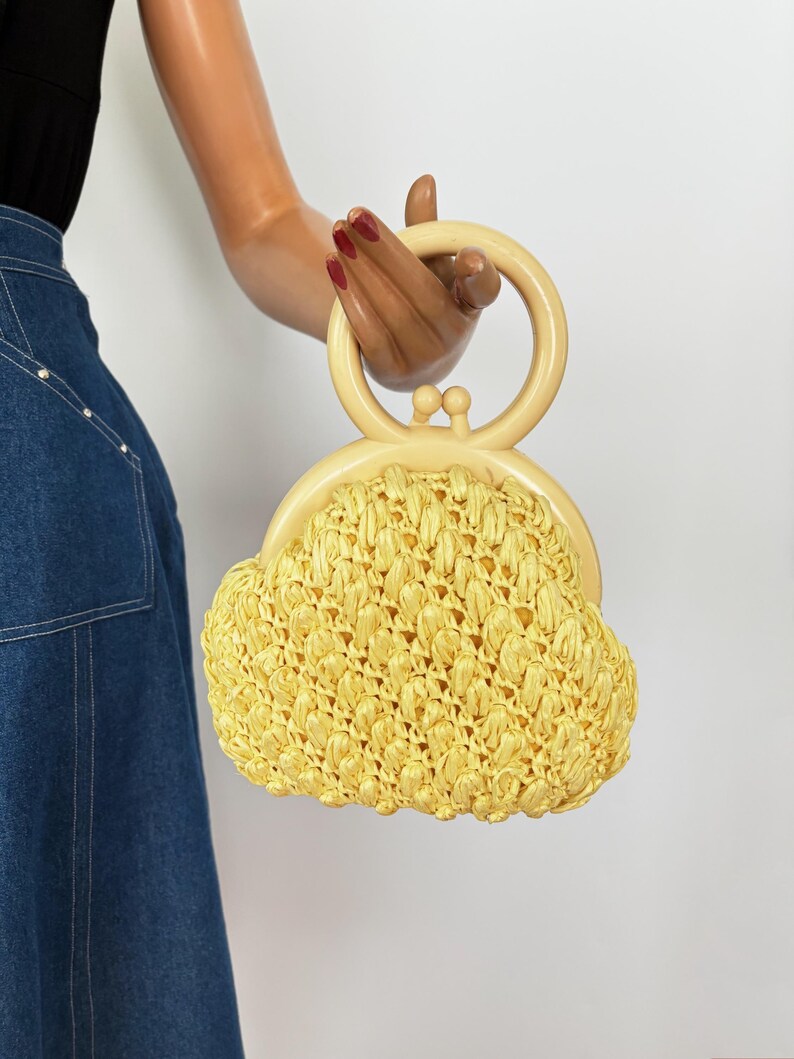 May include: A vintage yellow raffia handbag with a cream-colored handle and clasp. The bag has a rounded shape and a woven texture. The handle is a circular design. The bag is held by a mannequin.
