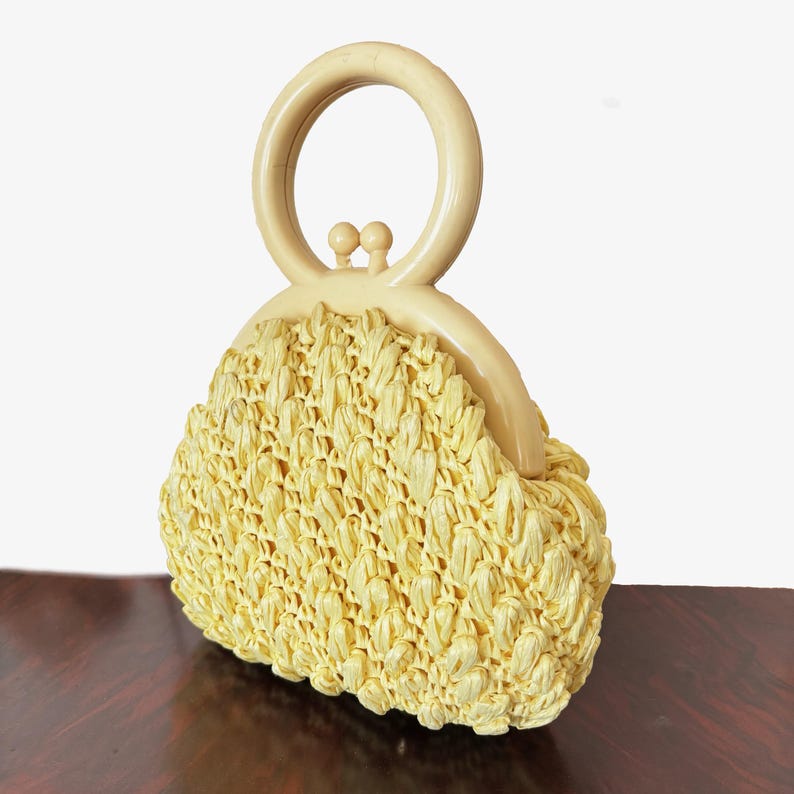 May include: A vintage, pale yellow handbag with a woven raffia body and a cream-coloured handle. The bag has a rounded shape and a clasp closure. The handle is a large, circular design. The bag is a classic accessory.