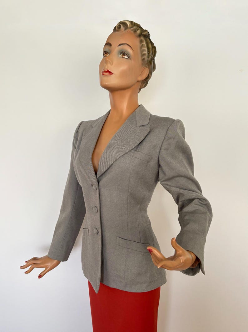 Puede incluir: Una blazer gris claro con corte entallado, con solapas de muesca y tres botones. La blazer tiene un bolsillo en el pecho y se combina con una falda roja. El maniqu&iacute; est&aacute; colocado sobre un fondo blanco.