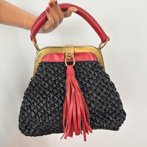 Op de afbeelding: Een handtas met een zwart gevlochten strooien lichaam, een rode lederen bovenrand en een bijpassend rood lederen handvat. De tas heeft een messingkleurig metalen frame met een centrale sluiting en een decoratieve rode lederen kwast.