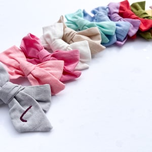 May include: A collection of colorful fabric hair bows arranged in a semi-circle. The bows are made of a soft, textured fabric and are tied with a simple knot. The bows are in various colors, including pink, blue, green, yellow, and red.
