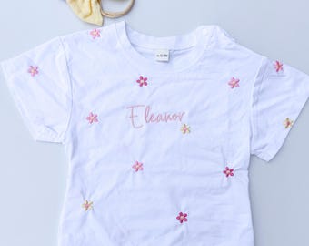 May include: White baby onesie with short sleeves and pink embroidered flowers. The onesie has the name "Eleanor" embroidered on the front in pink.
