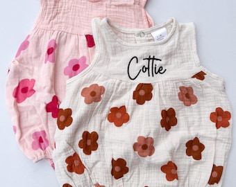 May include: Two sleeveless baby onesies with floral prints. The onesie on top is pink with pink flowers. The onesie on the bottom is beige with brown flowers and the name "Cottie" embroidered on the front.