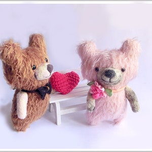 以下が含まれることがあります： 茶色とピンクのぬいぐるみクマ2体が白いベンチに座っています。茶色のクマは赤いハートを持っていて、ピンクのクマはピンクの花を持っています。