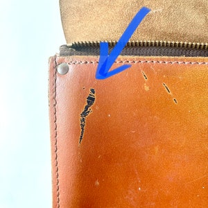 May include: Close-up of a brown leather bag with a zipper and a brass rivet. The leather has a few scratches and scuffs.