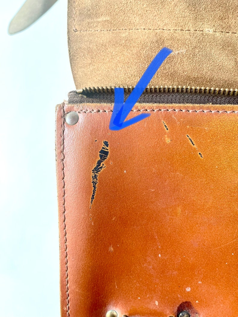 May include: Close-up of a brown leather bag with a zipper and a brass rivet. The leather has a few scratches and scuffs.