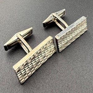 Könnte beinhalten: Ein Paar silberne Manschettenknöpfe mit geometrischem Muster. Die Manschettenknöpfe haben eine strukturierte Oberfläche und eine polierte Oberfläche.