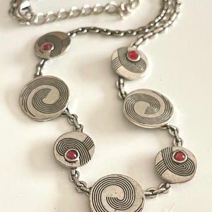 Puede incluir: Collar plateado con colgantes circulares con diseños en espiral y lineales. Cada colgante está conectado por una cadena y acentuado con una pequeña gema roja redonda. El collar tiene un cierre de mosquetón.