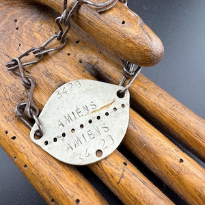 Könnte beinhalten: Ein antikes Metall-Hundemarke mit Kette, mit der Aufschrift "3420 AMIENS AMIENS 34 20". Die ovale Marke hat mehrere kleine Löcher. Kette und Marke weisen ein verwittertes, gealtertes Aussehen auf.