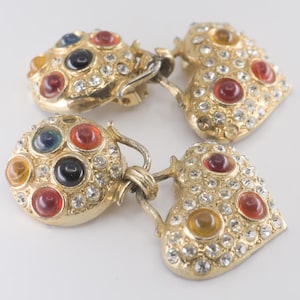 May include: Vintage gold-coloured clip-on earrings. One earring is round with colourful cabochons and clear rhinestones. The other earring is heart-shaped with red and yellow cabochons and clear rhinestones. The earrings are connected by a metal chain.
