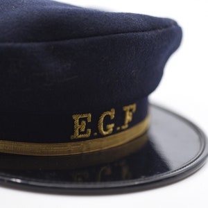 Può includere: Un berretto blu scuro con una fascia dorata e le lettere "E.G.F." ricamate in oro.