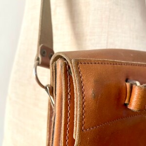 May include: A brown leather satchel bag with a long strap and a silver buckle. The bag has a flap closure and is made of high-quality leather.