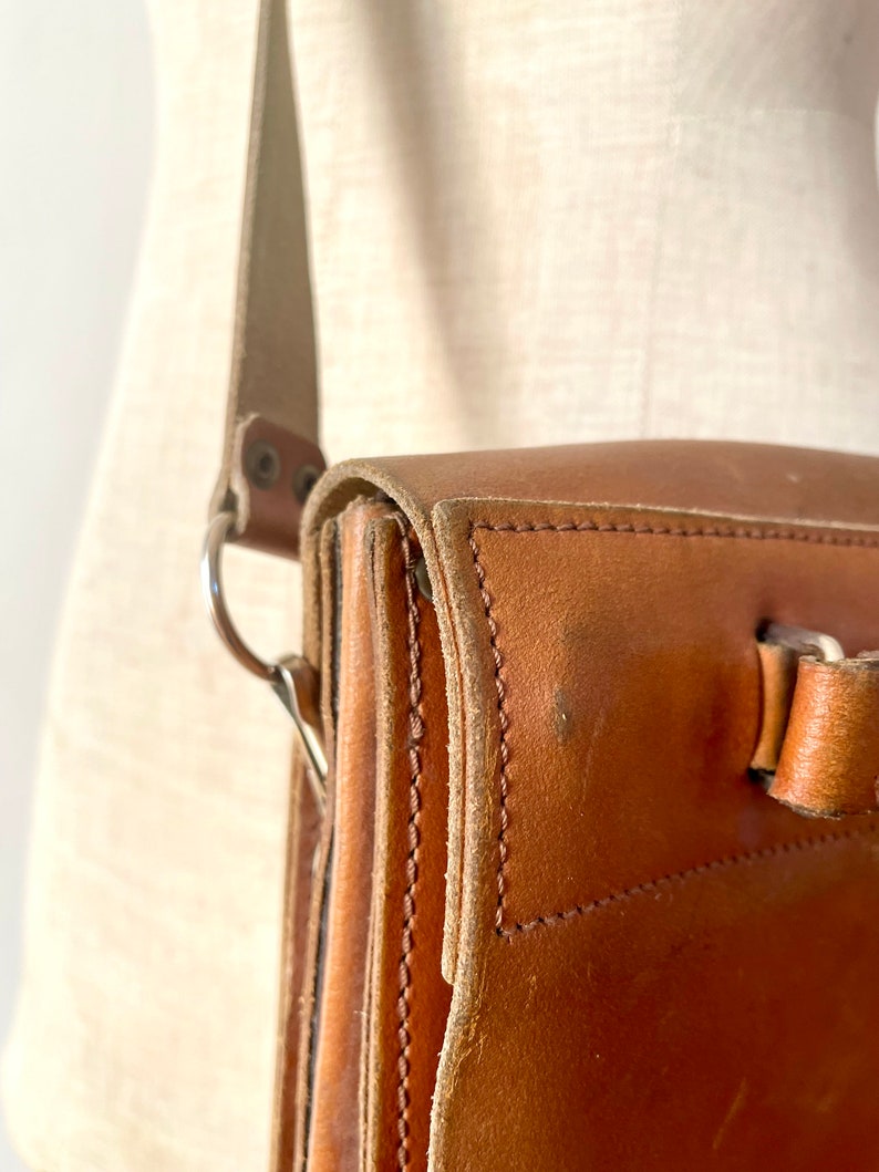 May include: A brown leather satchel bag with a long strap and a silver buckle. The bag has a flap closure and is made of high-quality leather.