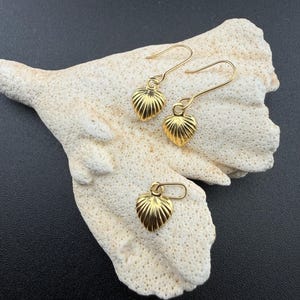 May include: Gold-coloured heart-shaped earrings with a textured, ridged design. The earrings are displayed on a piece of white coral against a black background. The image shows two earrings and a single charm.