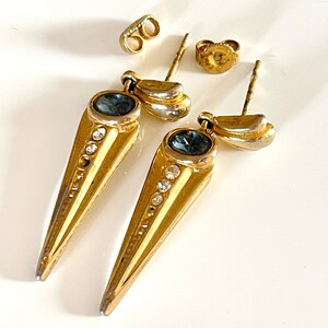 Puede incluir: Un par de pendientes de tono dorado con una piedra azul y brillantes transparentes. Los pendientes tienen forma de cono puntiagudo.
