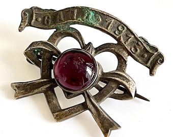 May include: Antique silver-toned brooch with a large, round, deep red gemstone at its center. The brooch features a ribbon design with the inscription "GAIL 1913". The pin is a vintage accessory.