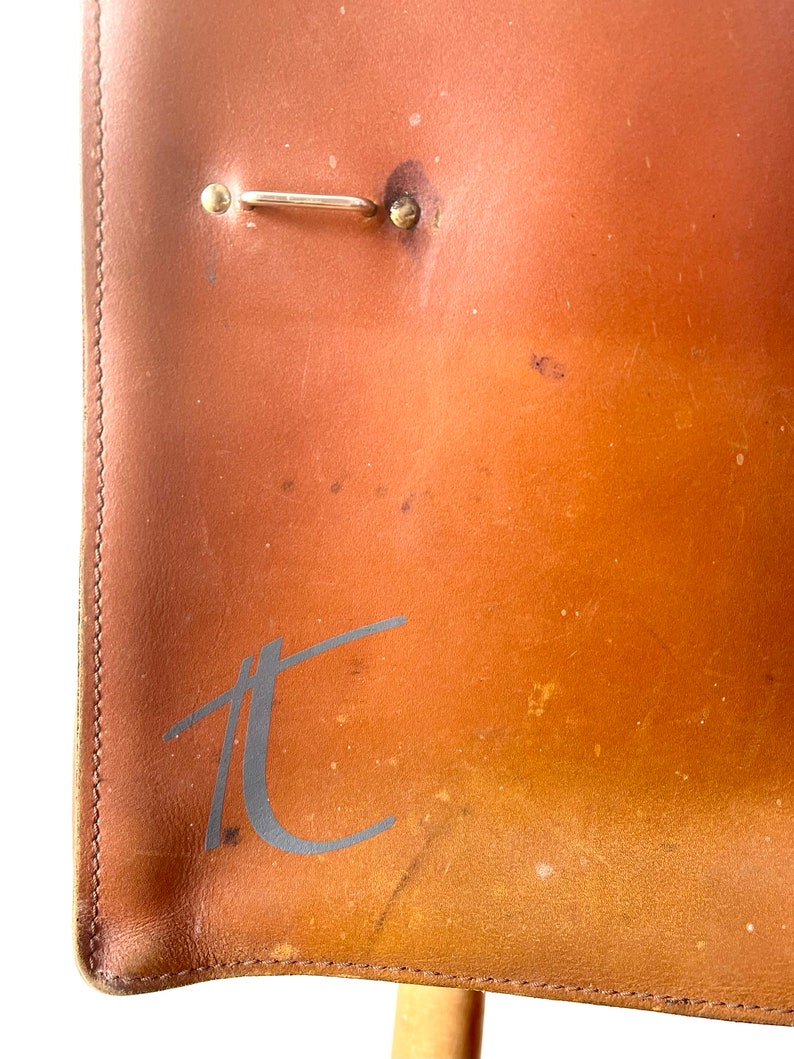 May include: Close-up of a brown leather chair with a metal hook and the letter "T" embossed on the back.