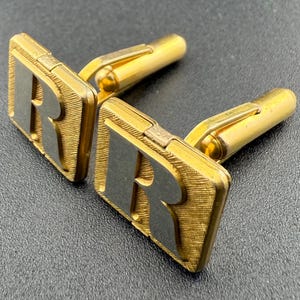 May include: A pair of gold cufflinks with the letter "R" engraved on each. The cufflinks have a textured surface and a polished finish.