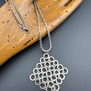 Peut inclure: Collier argenté avec un pendentif géométrique composé de cercles entrelacés. La chaîne est de conception simple, et le pendentif est en forme de losange. Le collier est présenté sur une main en bois sur fond sombre.