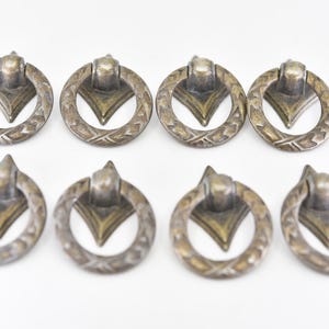 Peut inclure: Huit boutons de cabinet en laiton antique avec une bague circulaire et un motif triangulaire au centre. Les boutons ont un motif décoratif et orné autour de la bague.