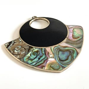 May include: A silver pendant with a black center circle and a surrounding design of iridescent abalone shell. The pendant has a circular opening at the top for a chain.