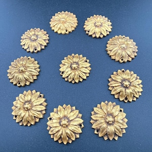 Op de afbeelding: Tien goudkleurige, zonnebloemvormige decoratieve versieringen. Elk stuk heeft gedetailleerde bloemblaadjes en kleine gaatjes voor bevestiging. De versieringen zijn in een cirkelvormig patroon op een donkere achtergrond gerangschikt.