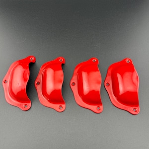 Puede incluir: Cuatro tiradores de cajón tipo concha en metal rojo brillante dispuestos en fila horizontal. Cada tirador presenta una zona de agarre semicircular lisa y una brida de montaje plana con tres pequeños orificios para tornillos.