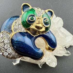Puede incluir: Un broche de panda en tono dorado con esmalte azul y verde y pedrería transparente. El panda está sentado con las patas delante.