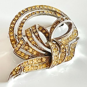 May include: A silver and gold tone brooch with a bow design. The bow is made of small, clear rhinestones.