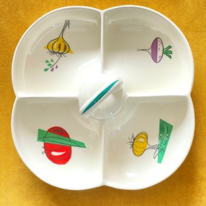Puede incluir: Plato de servir de cerámica blanca con cuatro secciones, cada una con una ilustración de verdura diferente: una cebolla amarilla, un tomate rojo, un nabo morado y un ajo amarillo. El plato tiene un asa verde.