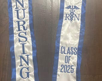 May include: A white and blue graduation sash with the word "NURSING" embroidered in blue. The sash also features "BSN R.N." and "CLASS OF 2025". The sash is made of a satin-like material.