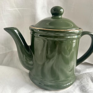 Może przedstawiać: Biały ceramiczny kubek z czarnym uchwytem i napisem "But First, Coffee" w stylu odręcznym. Kubek stoi na białej powierzchni. Kubek ma około 10 cm wysokości.