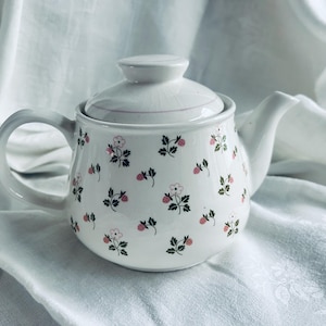 Könnte beinhalten: Weiße Keramik-Teekanne mit Deckel, Henkel und Ausguss. Die Teekanne ist mit einem Muster aus kleinen rosa Blüten und grünen Blättern verziert. Die Teekanne steht auf einem weißen Tuch.