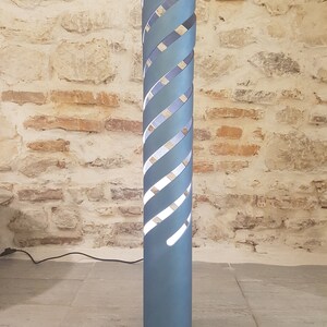 Op de afbeelding: Een hoge, lichtblauwe vloerlamp met een spiraalvormig ontwerp. De lamp heeft een wit licht dat schijnt door de uitsparingen in de spiraal.