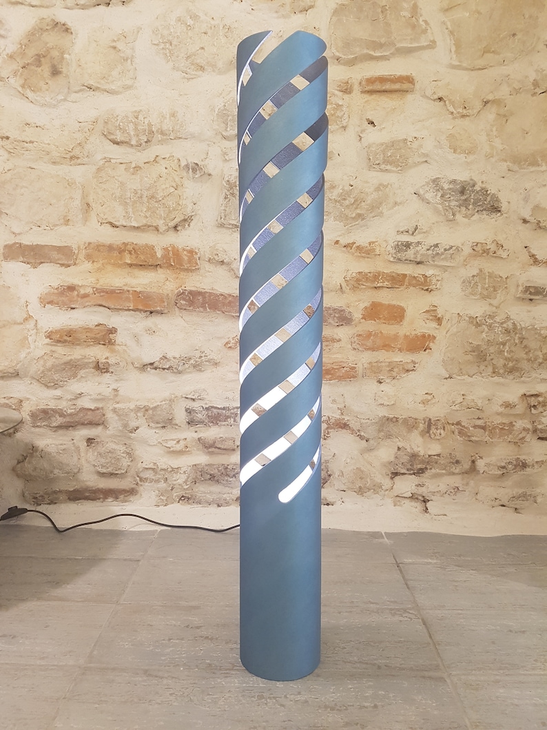 Op de afbeelding: Een hoge, lichtblauwe vloerlamp met een spiraalvormig ontwerp. De lamp heeft een wit licht dat schijnt door de uitsparingen in de spiraal.