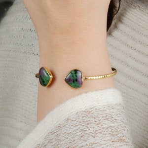 Può includere: Un braccialetto rigido dorato con due pietre a forma di cuore verde e rosa.
