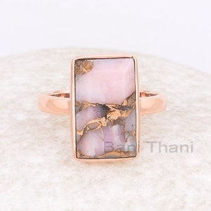 Puede incluir: Un anillo de oro rosa con una piedra rectangular rosa y dorada. El anillo está colocado sobre una roca blanca.