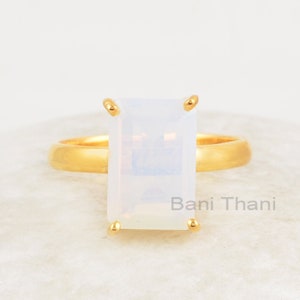 May include: A gold ring with a white rectangular gemstone. The ring is on a white surface. "Bani Thani" is written on the surface.