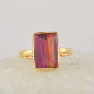 Könnte beinhalten: Ein goldener Ring mit einem rechteckigen, schillernden Edelstein. Der Edelstein hat Farbtöne von Rot, Lila und Orange.