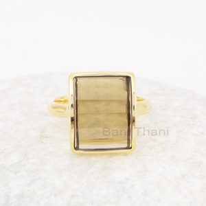Może przedstawiać: Złoty pierścionek z prostokątnym kamieniem z dymnego kwarcu. Pierścionek leży na białym tle.