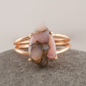Puede incluir: Un anillo de oro rosa con una piedra preciosa rosa y marrón en forma de lágrima. El anillo tiene un diseño de doble banda.