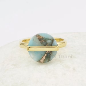 Puede incluir: Anillo de oro con una piedra ovalada de color azul claro y oro. El anillo tiene una banda de oro con una barra horizontal de oro en el centro de la piedra.