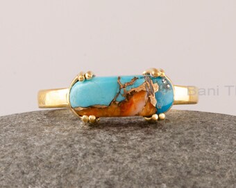 May include: A gold ring with a rectangular turquoise stone set in the center. The stone has a blue and orange marbled pattern. The ring is on a gray rock.