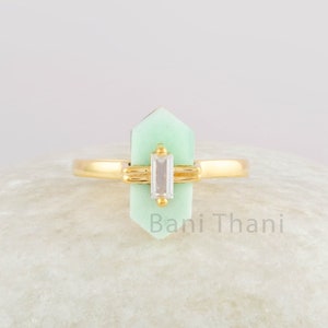 Peut inclure: Une bague en or avec une pierre précieuse vert clair et une pierre rectangulaire transparente. La bague est posée sur une pierre blanche. Le texte "Bani Thani" est inscrit sur la pierre.
