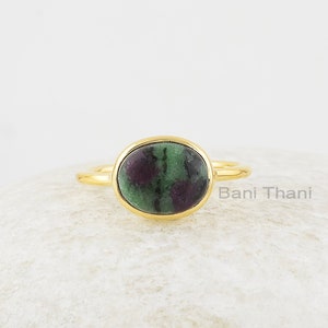 May include: Gold ring featuring a green and purple gemstone. The ring is displayed on a white stone.