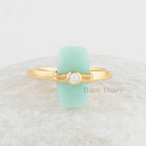 May include: A gold ring featuring a light green stone and a small clear stone in the centre. The ring is displayed on a grey stone background.