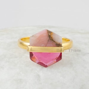 Può includere: Anello in oro con una pietra esagonale rosa e bianca. L'anello è posto su una superficie bianca.